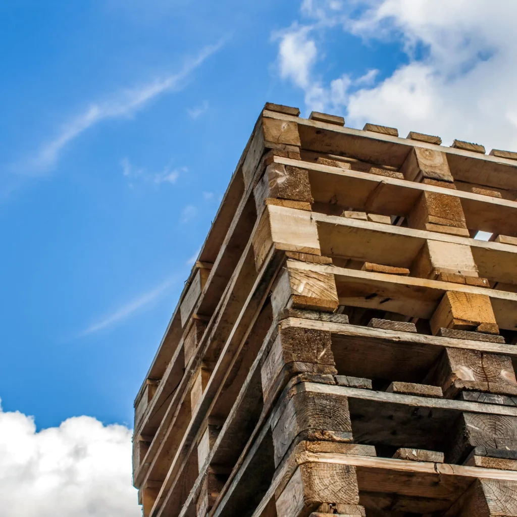 Stos drewnianych palet ułożonych na zewnątrz, na tle błękitnego nieba z białymi chmurami. Palety są przygotowane do dalszego wykorzystania lub transportu.