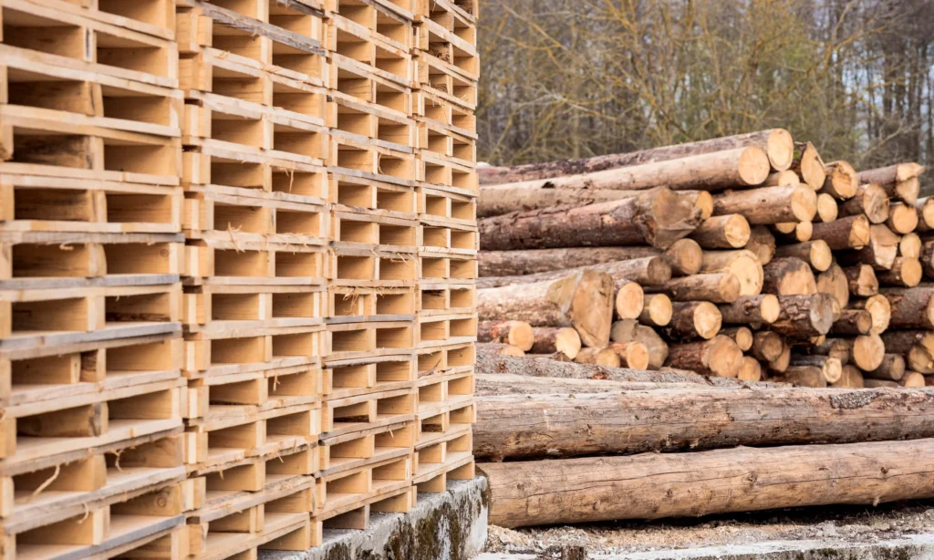 Stosy drewnianych palet gotowych do użycia oraz pnie drzew przygotowane do obróbki w tle. Palety są kluczowe w logistyce i magazynowaniu towarów.