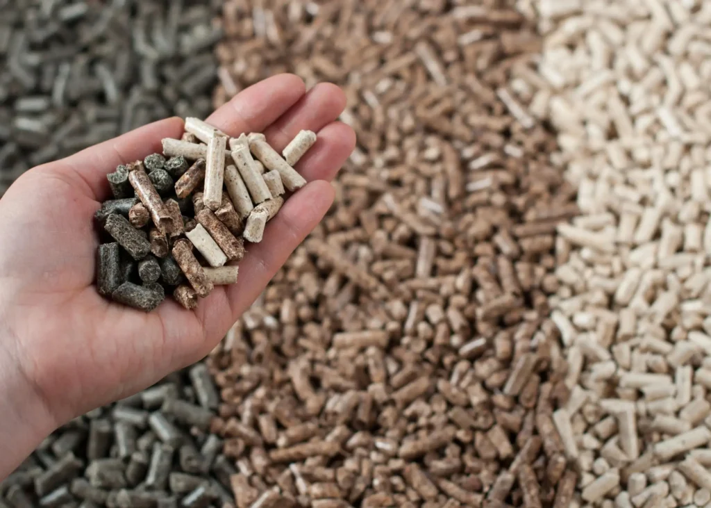 Dłoń trzymająca różnokolorowy pellet opałowy na tle większej ilości pelletu, symbolizująca różnorodność materiałów paliwowych.