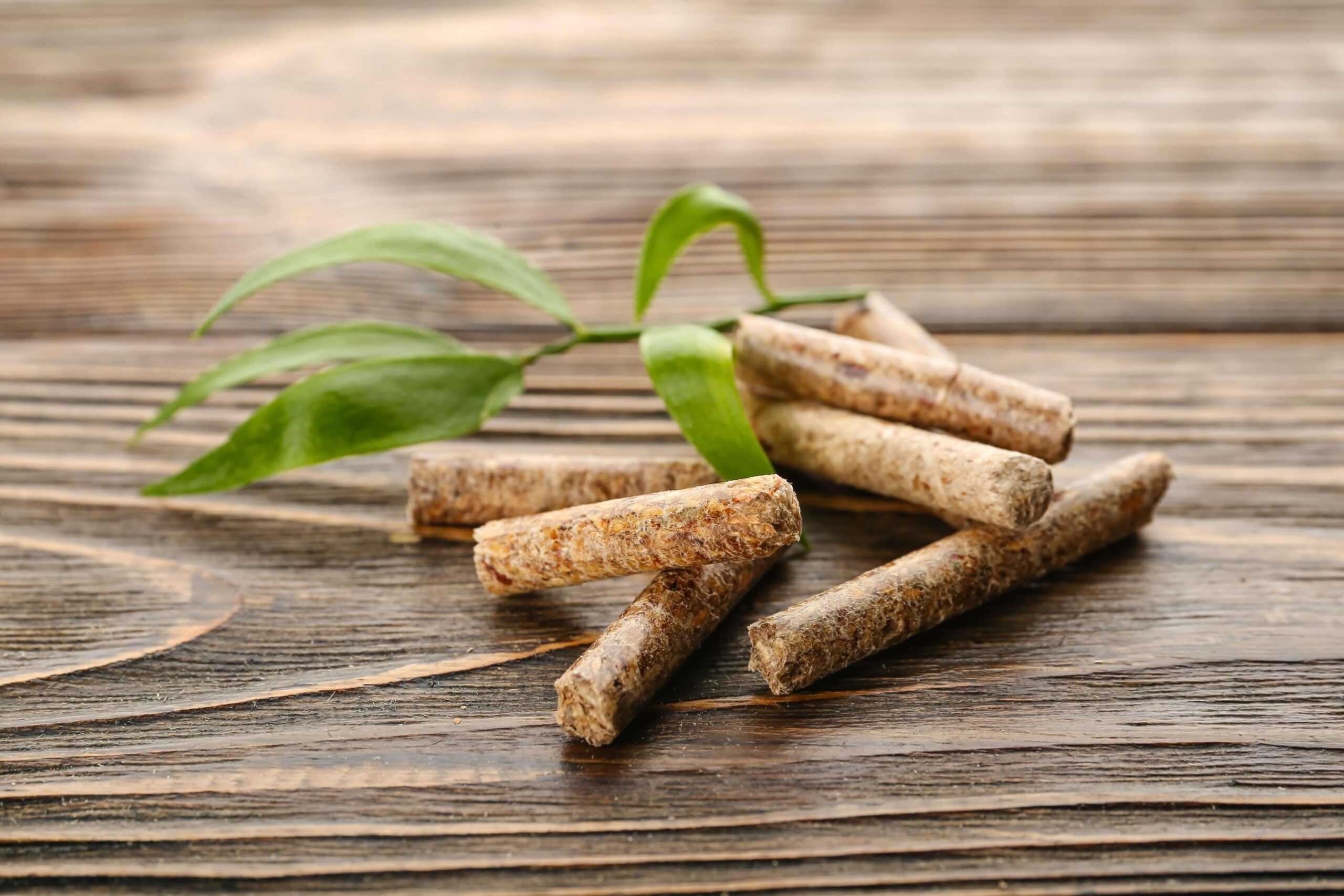 Zdjęcie przedstawia drewniane pellety ułożone na starej desce z zielonymi liśćmi w tle, co podkreśla ich ekologiczny charakter.