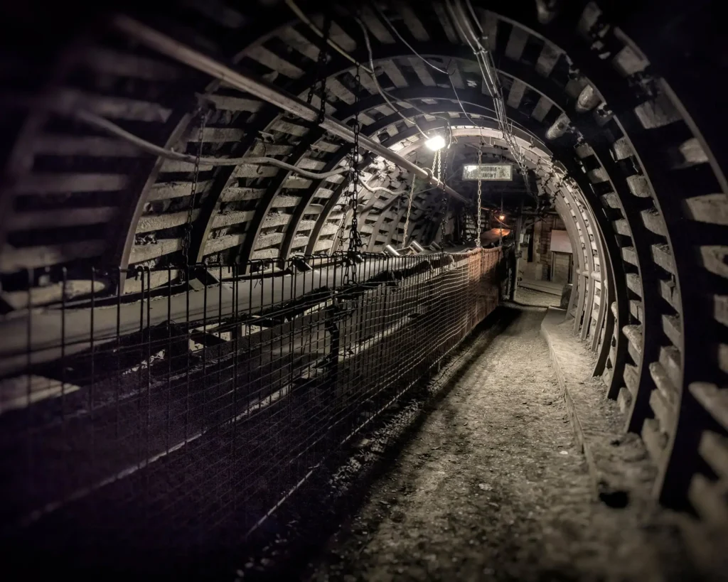 Widok z wnętrza tunelu kopalni węgla kamiennego, otoczonego metalowymi i betonowymi wzmocnieniami, z oświetleniem technicznym.