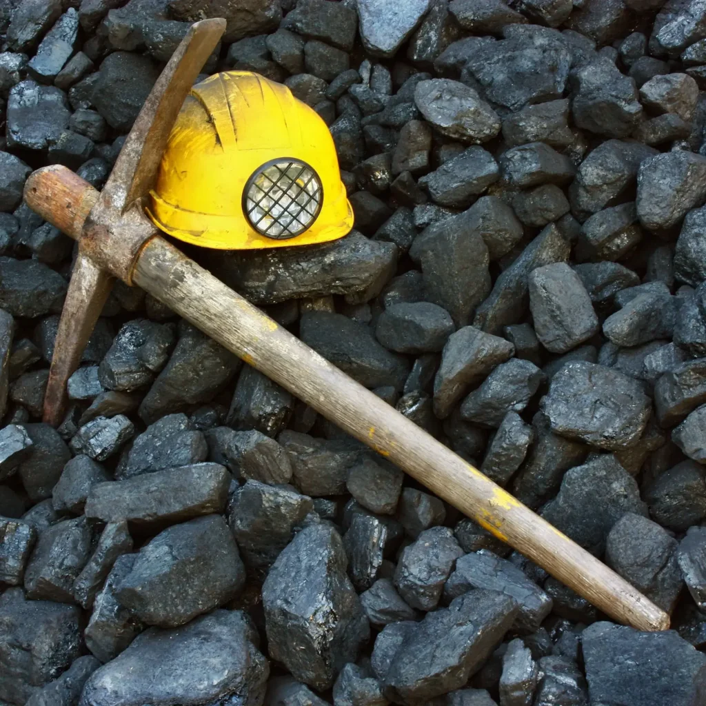 Żółty kask ochronny z lampką górniczą oraz stary kilof leżące na stosie węgla orzech, symbolizujące pracę w kopalni.