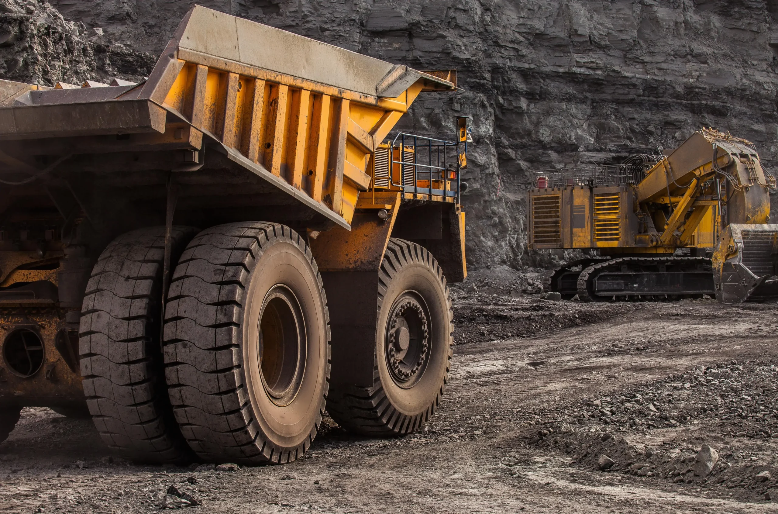 Duża żółta wywrotka i koparka w kopalni węgla, na tle ciemnoszarej ściany skalnej.
